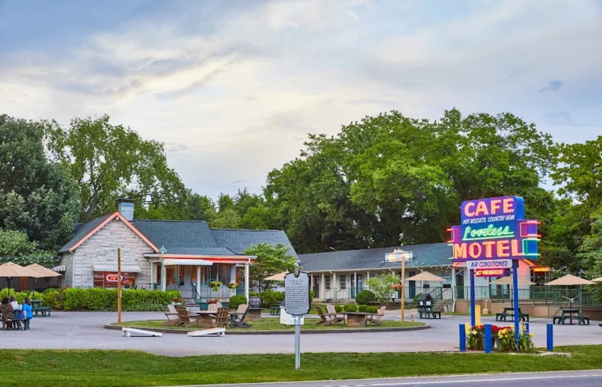A small roadside motel and café with vintage neon signs, outdoor seating, and picnic tables, surrounded by trees and a clear, partly cloudy sky in the background.