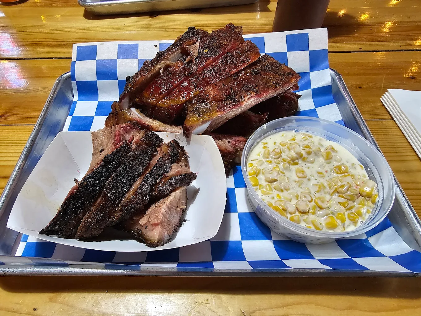 A metal tray with barbecued ribs, sliced brisket, and a side of creamed corn in a plastic container, all on blue-and-white checkered paper atop a wooden table.