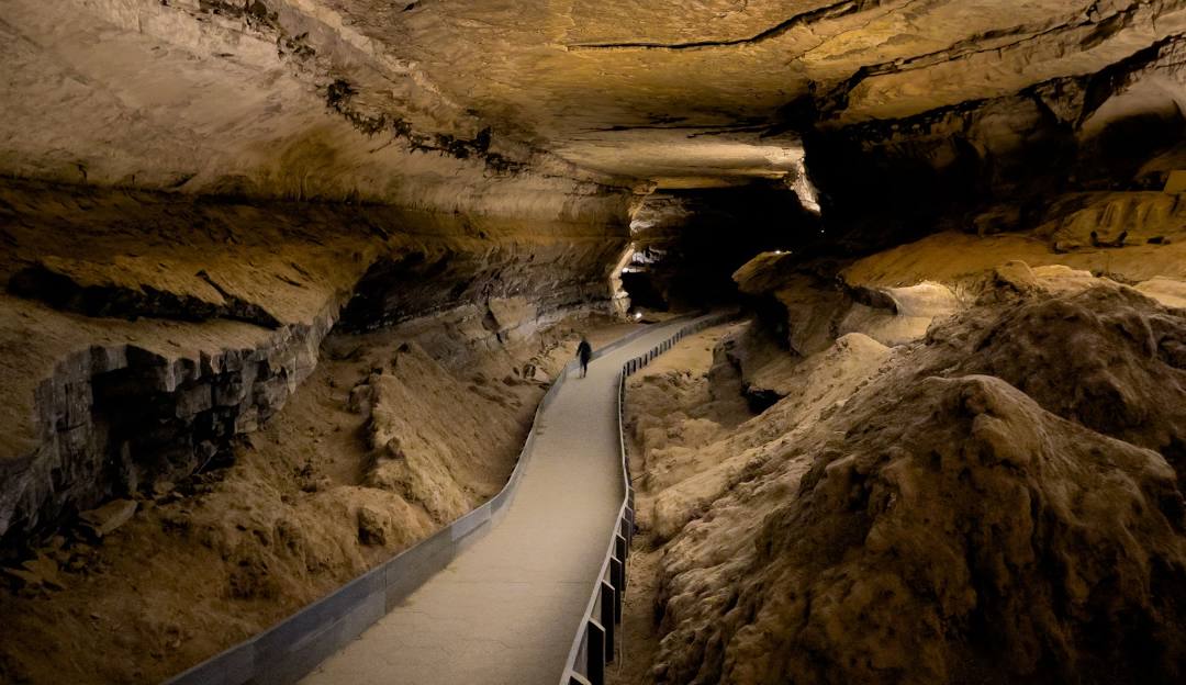 A person walks along a paved path with railings inside a large, dimly lit cave with rough, rocky walls and ceiling. Southern travel hidden gems. 