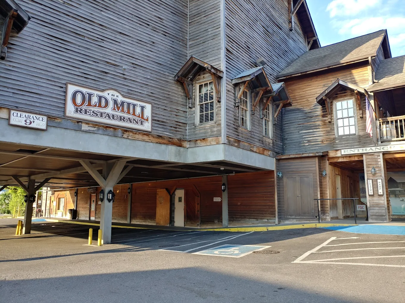 The exterior of The Old Mill Restaurant, a large wooden building with rustic architecture, a covered parking area, and accessible parking spaces near the entrance. Signs indicate the entrance and a 9-foot clearance for vehicles.