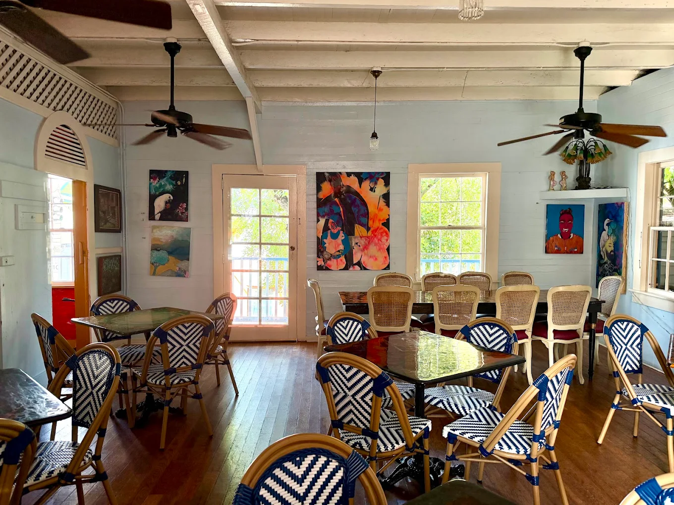 A bright, airy café with wooden floors, blue and white woven chairs, glass-top tables, colorful paintings on the walls, ceiling fans, and large windows letting in natural light.
