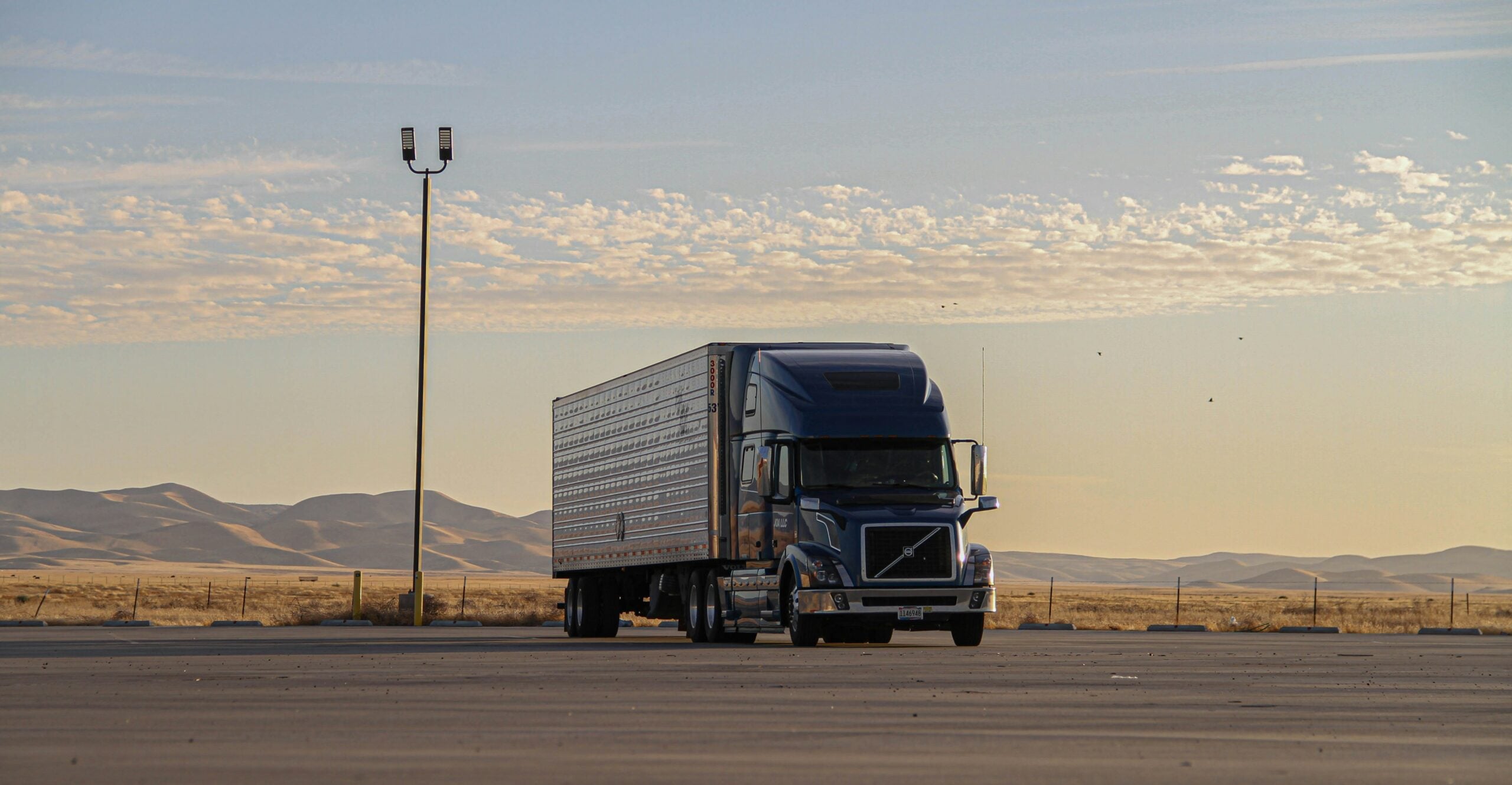 A blue semi-truck parked in an empty lot with mountains and a partly cloudy sky in the background during daylight.