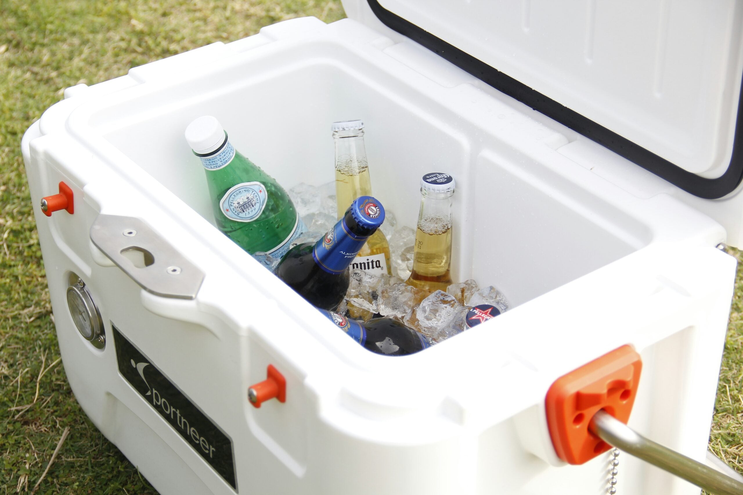 A white cooler filled with ice contains bottles of sparkling water, beer, and other beverages. The cooler is open and sitting on grass outdoors.