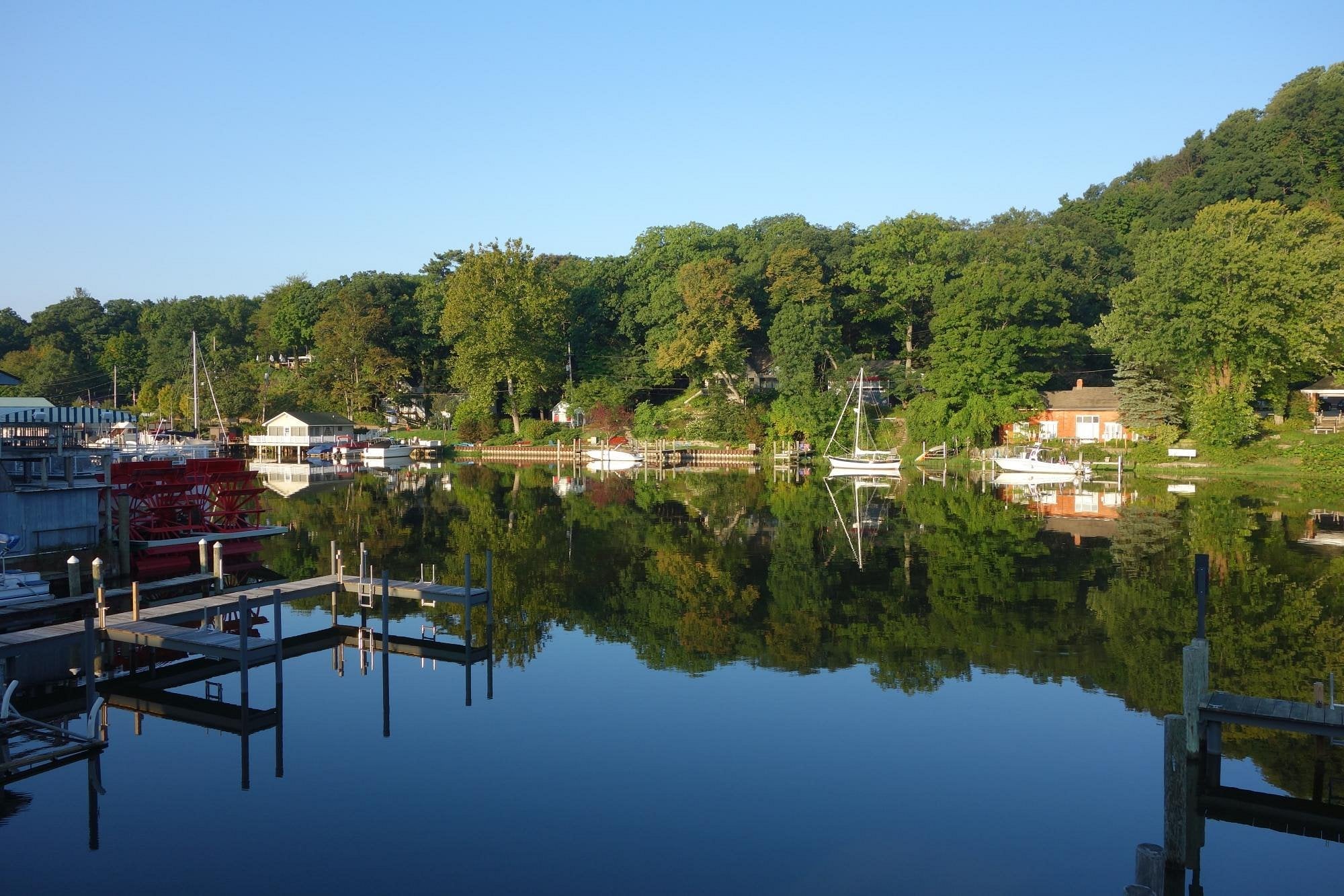 A calm marina with empty docks and boats moored along a tree-lined shore, all reflected clearly in the still water under a bright blue sky—a tranquil scene often found in unique American towns.