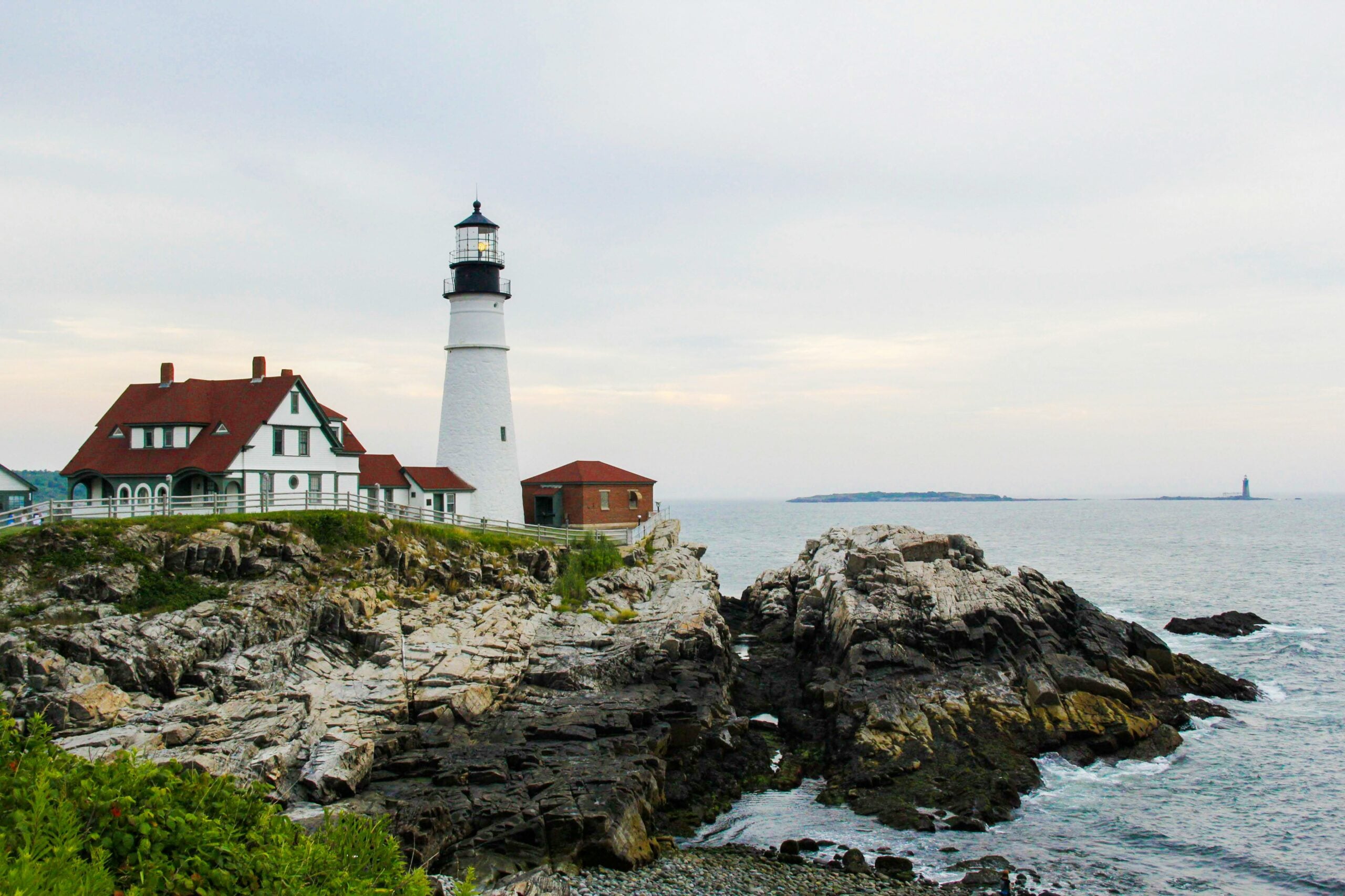 A white lighthouse with a black top stands on rocky cliffs by the ocean, next to a house with a red roof. The sky is cloudy and small islands are visible in the distance over the water.