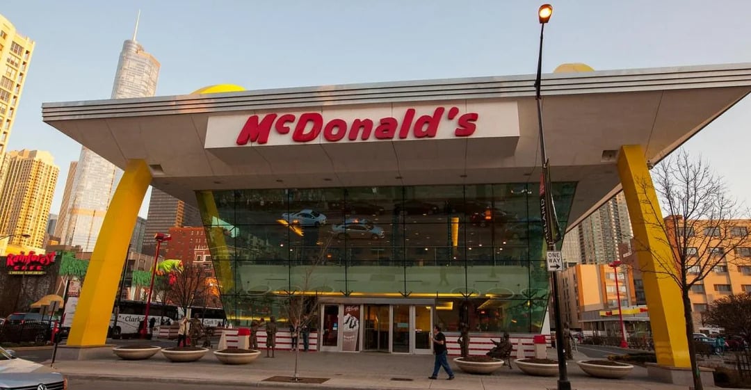 A modern McDonald’s restaurant with large glass windows and yellow arch supports, located in a city with tall buildings in the background—one of the coolest McDonald's locations. Several people are walking on the sidewalk outside.