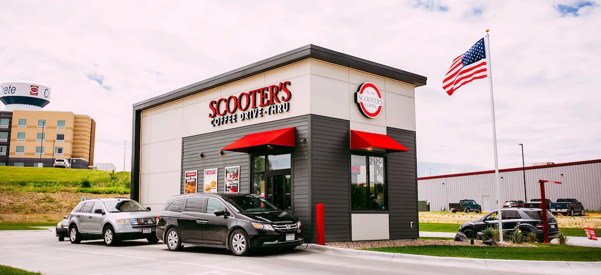 A Scooter's Coffee drive-thru location with two cars in line, a U.S. flag on a pole, and commercial buildings in the background under a partly cloudy sky.