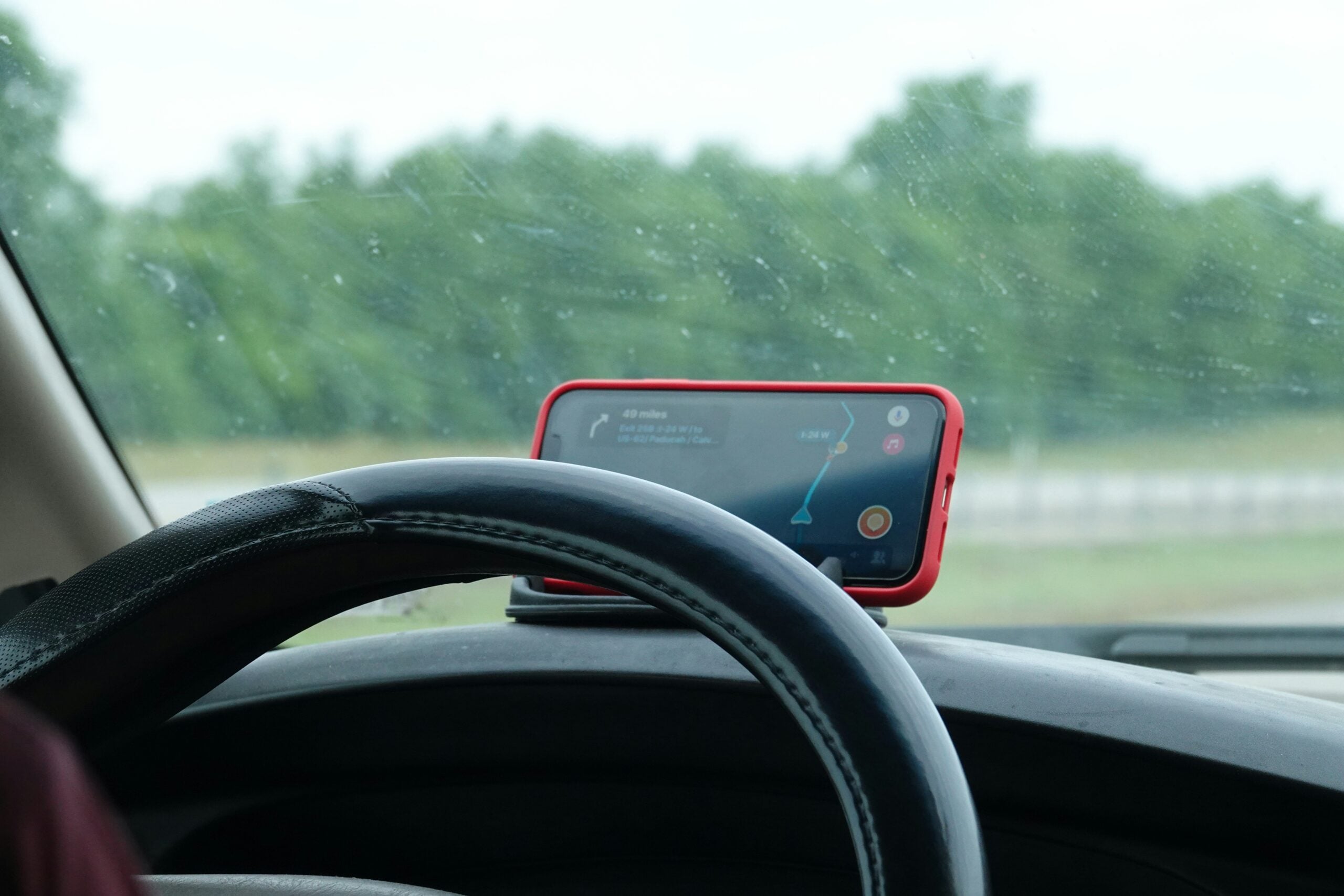 A smartphone with a red case displaying GPS navigation is mounted on a car dashboard behind the steering wheel, with a blurred view of trees and a road through the windshield.