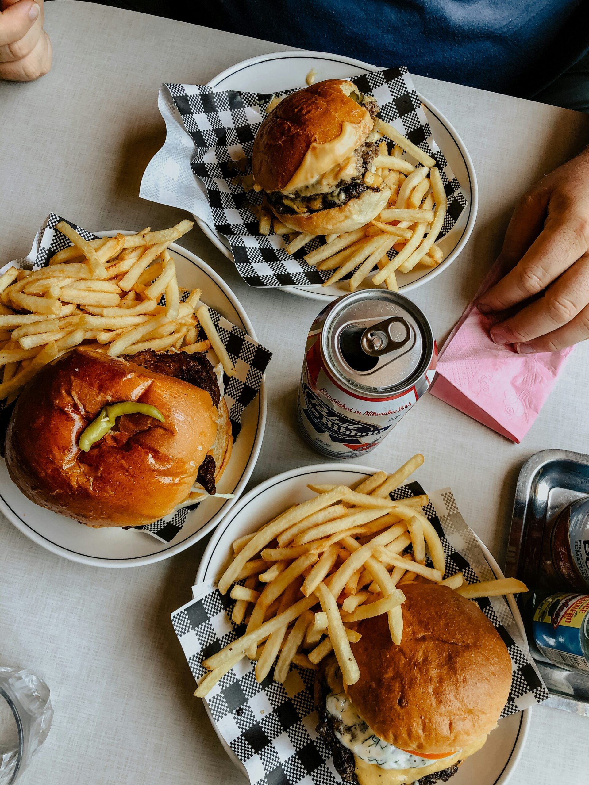 Three plates of cheeseburgers with fries are on a table, with a can of beer and a pink napkin.