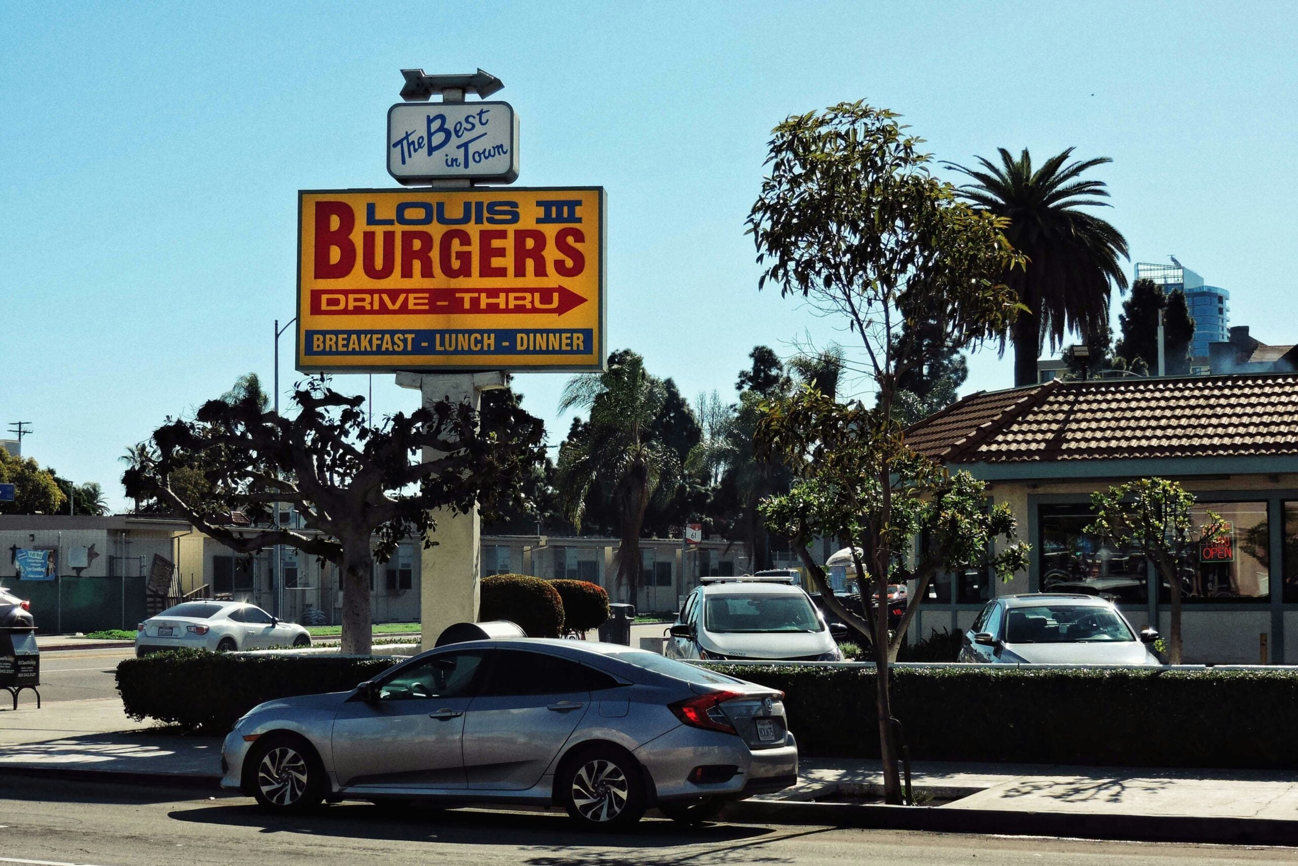 A silver car is parked in front of Louis Burgers III, a drive-thru restaurant with a large yellow and red sign advertising breakfast, lunch, and dinner. Palm trees and other cars are visible in the background.