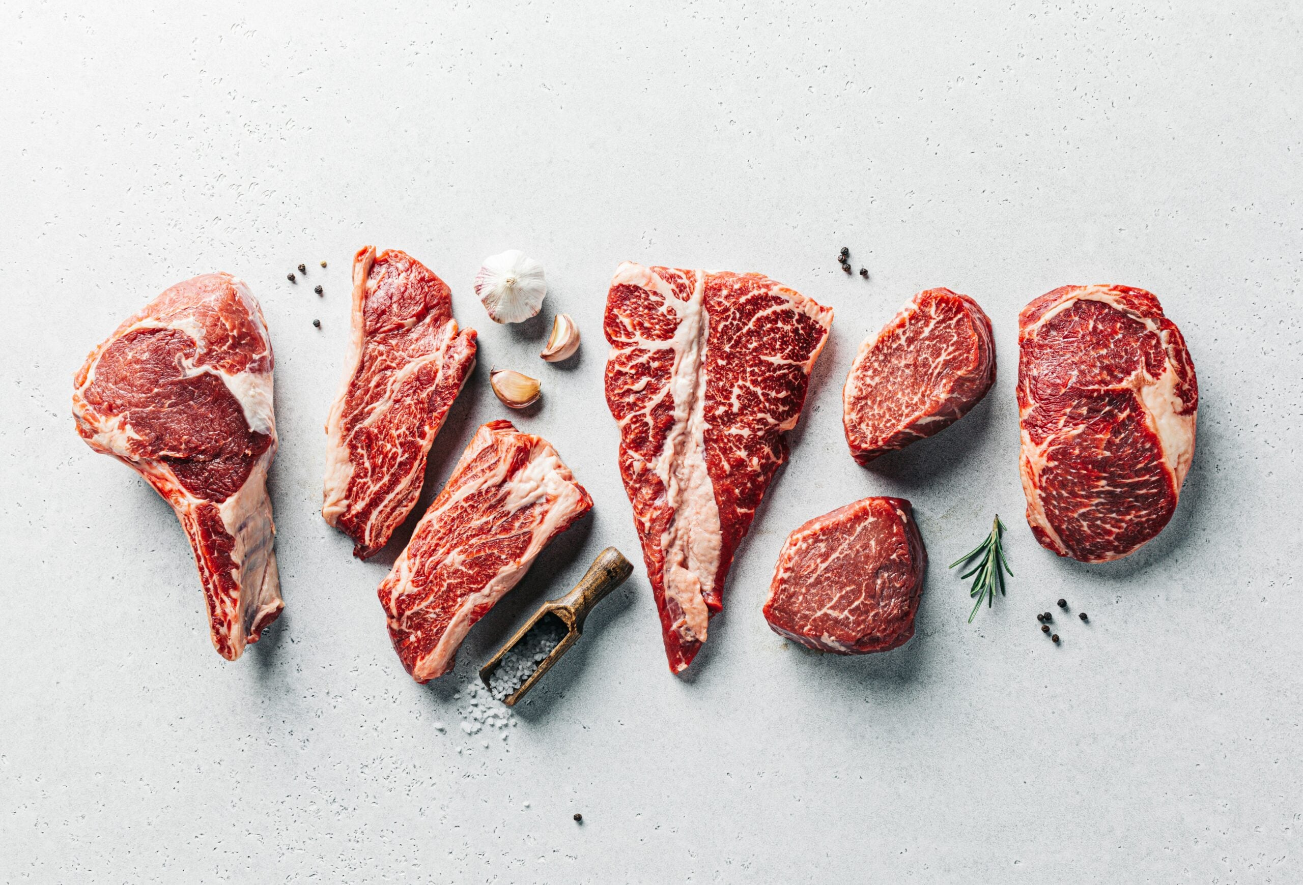 Seven different raw beef steaks arranged on a light surface, with scattered herbs, garlic cloves, peppercorns, coarse salt, and a small meat cleaver placed nearby.