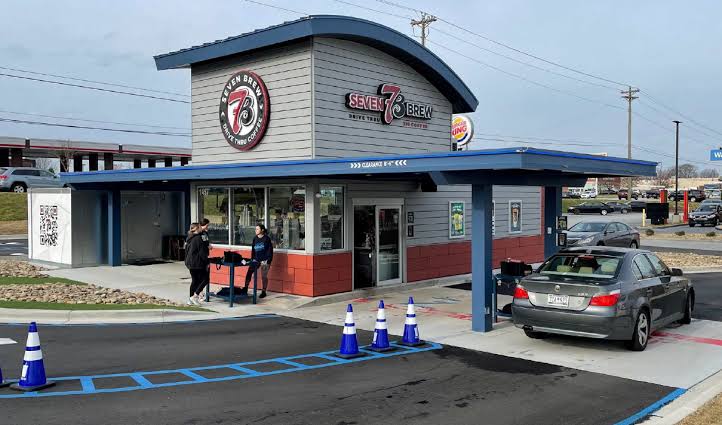 A drive-thru coffee stand named Seven Brew with cars in line, two people talking outside, and traffic cones near the entrance. The small building has a modern design and a large logo on the roof.