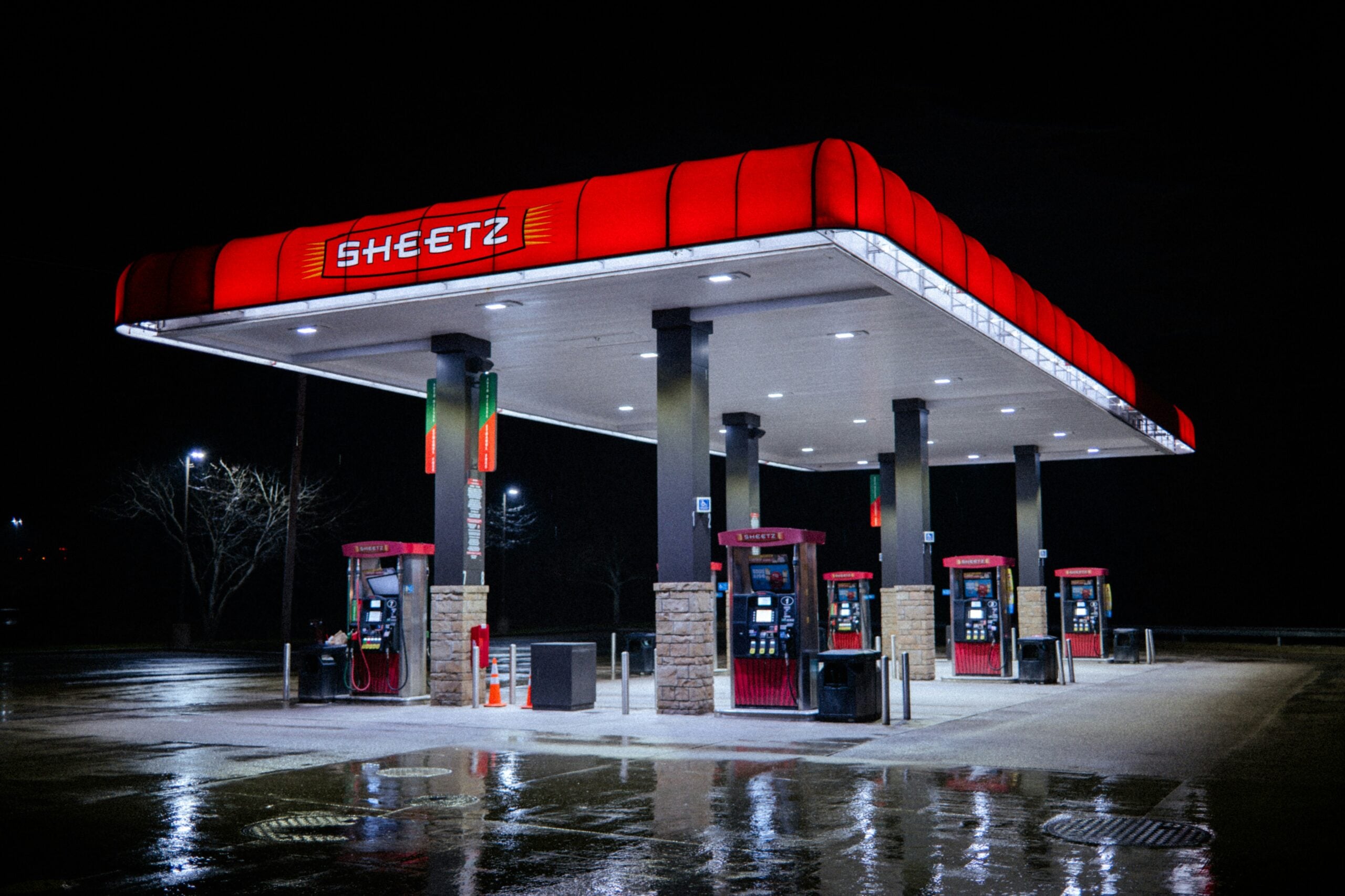 A brightly lit Sheetz gas station with red signage and four fuel pumps stands alone at night, surrounded by darkness and wet pavement reflecting the lights.
