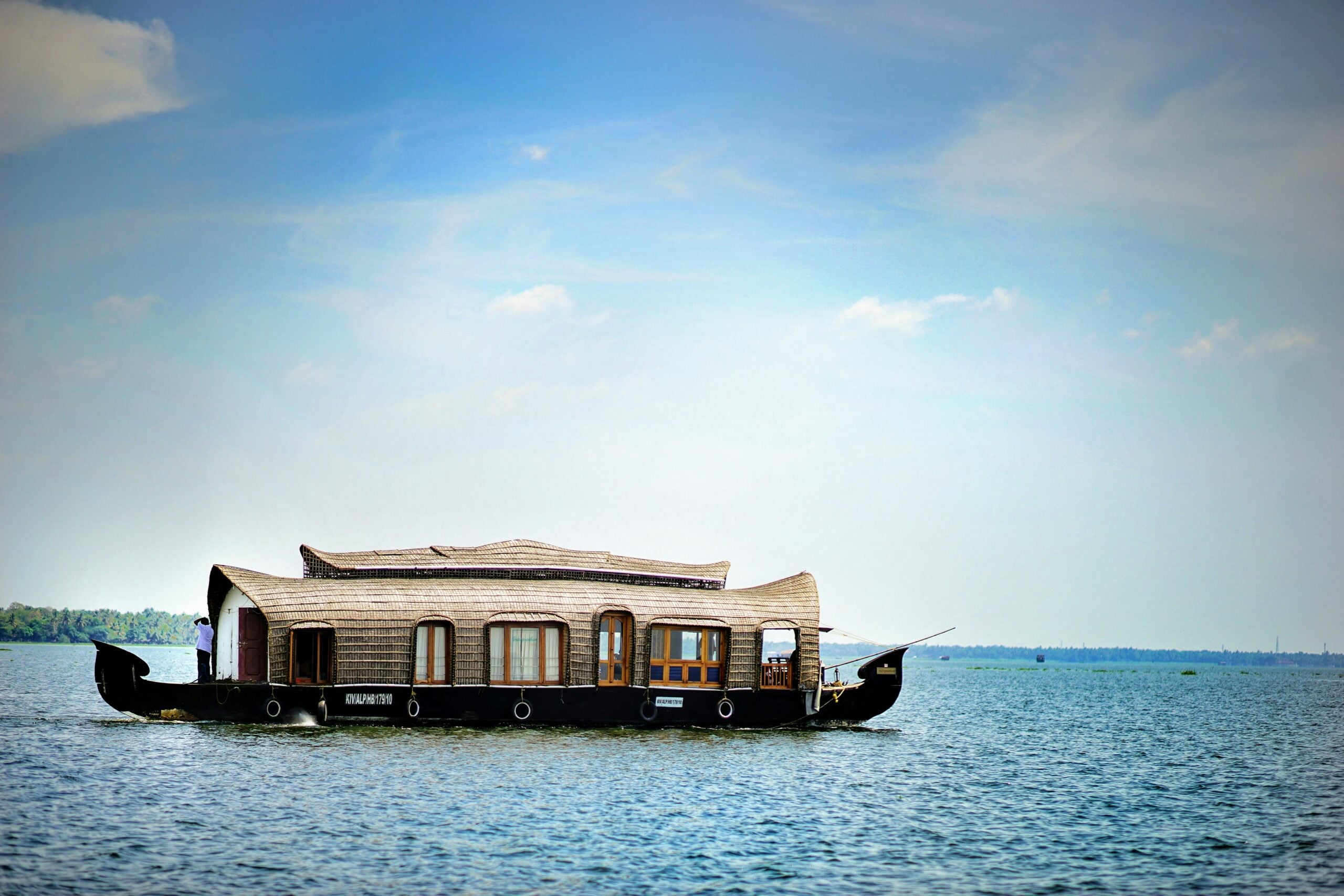 A traditional wooden houseboat with arched roof floats on calm blue water under a clear sky, with a distant green shoreline visible in the background.