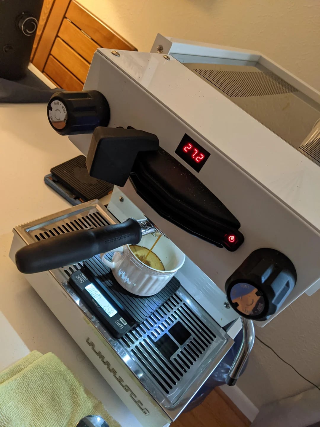 A white espresso machine with a portafilter is making coffee into a small white ramekin. The machine’s digital display reads "212," and various knobs and gauges are visible—perfect for fans of top coffee brands seeking precision brewing.