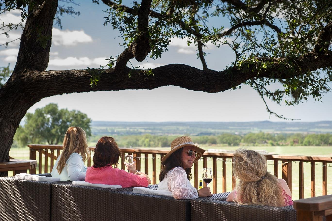 Four women sit on an outdoor patio under a large tree, enjoying wine and chatting while gazing out over fields and distant hills, soaking in the charm found in many unique American towns beneath a partly cloudy sky.