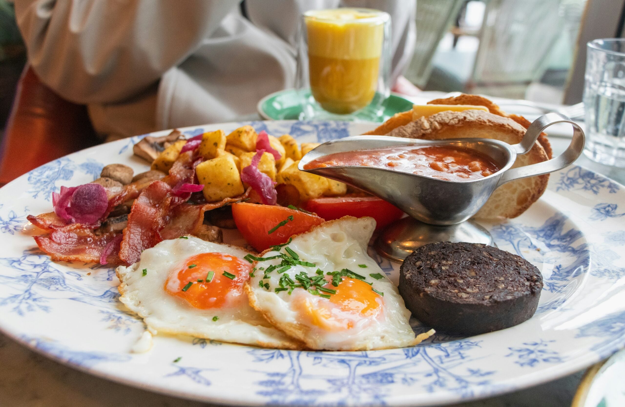 A plate with two sunny-side-up eggs, grilled tomato, bacon, sautéed potatoes with onions, mushrooms, a slice of black pudding, baked beans in a gravy boat, pita bread, and a glass of coffee in the background.