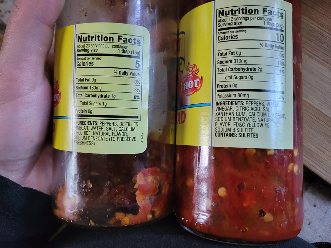 Close-up of two jars of hot sauce, showing their nutrition facts labels. The left label states 5 calories and 180mg sodium per serving. The right label shows 10 calories and 310mg sodium.