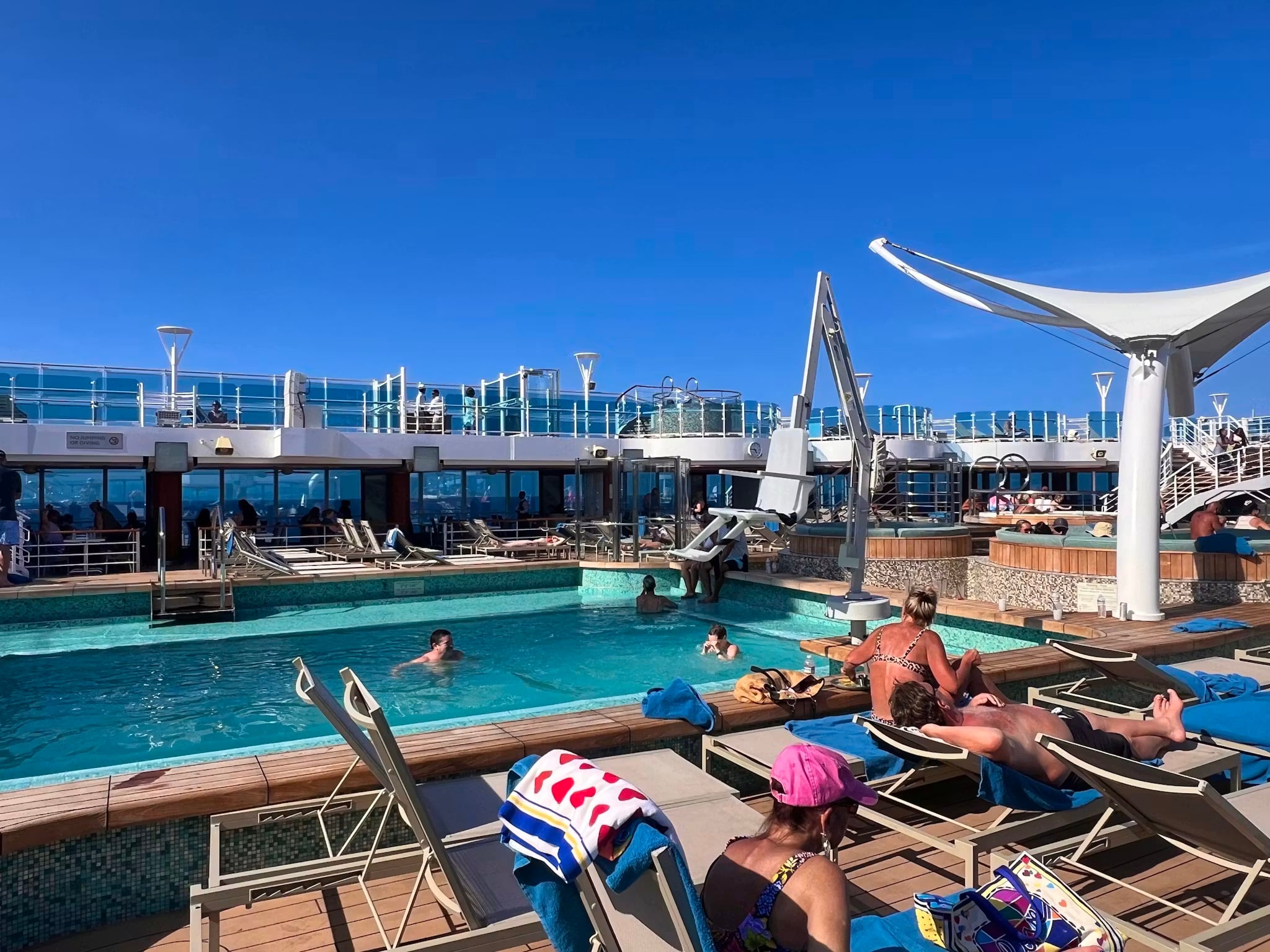 People relax on lounge chairs and swim in a pool under the clear blue sky aboard the Best Cruise Line 2026. Towels and bags are scattered around the deck, with the ship’s upper deck and railings in view.