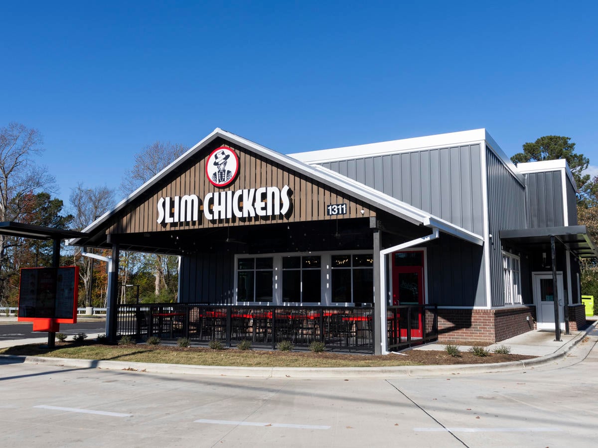 A Slim Chickens restaurant with a patio seating area, large sign above the entrance, and a drive-thru lane, set against a clear blue sky and surrounded by trees.