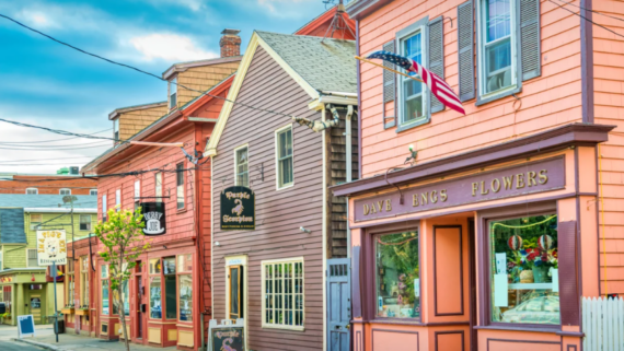 Colorful storefronts line a quaint street in a small town, reflecting the vibrant spirit of Small Business States. Among them is a flower shop with an American flag, lively window displays, and bright paint beneath a clear, partly cloudy sky.