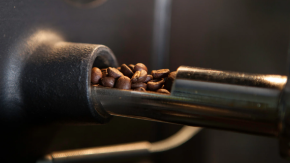 Close-up of dark roasted coffee beans being dispensed from a metal spout on a coffee roasting machine&mdash;a glimpse into the small luxuries Americans won't give up, with a beautifully blurred background.