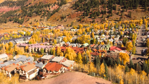 Aerial view of a unique American town with houses and buildings surrounded by trees with yellow autumn foliage, set against a backdrop of forested hills.