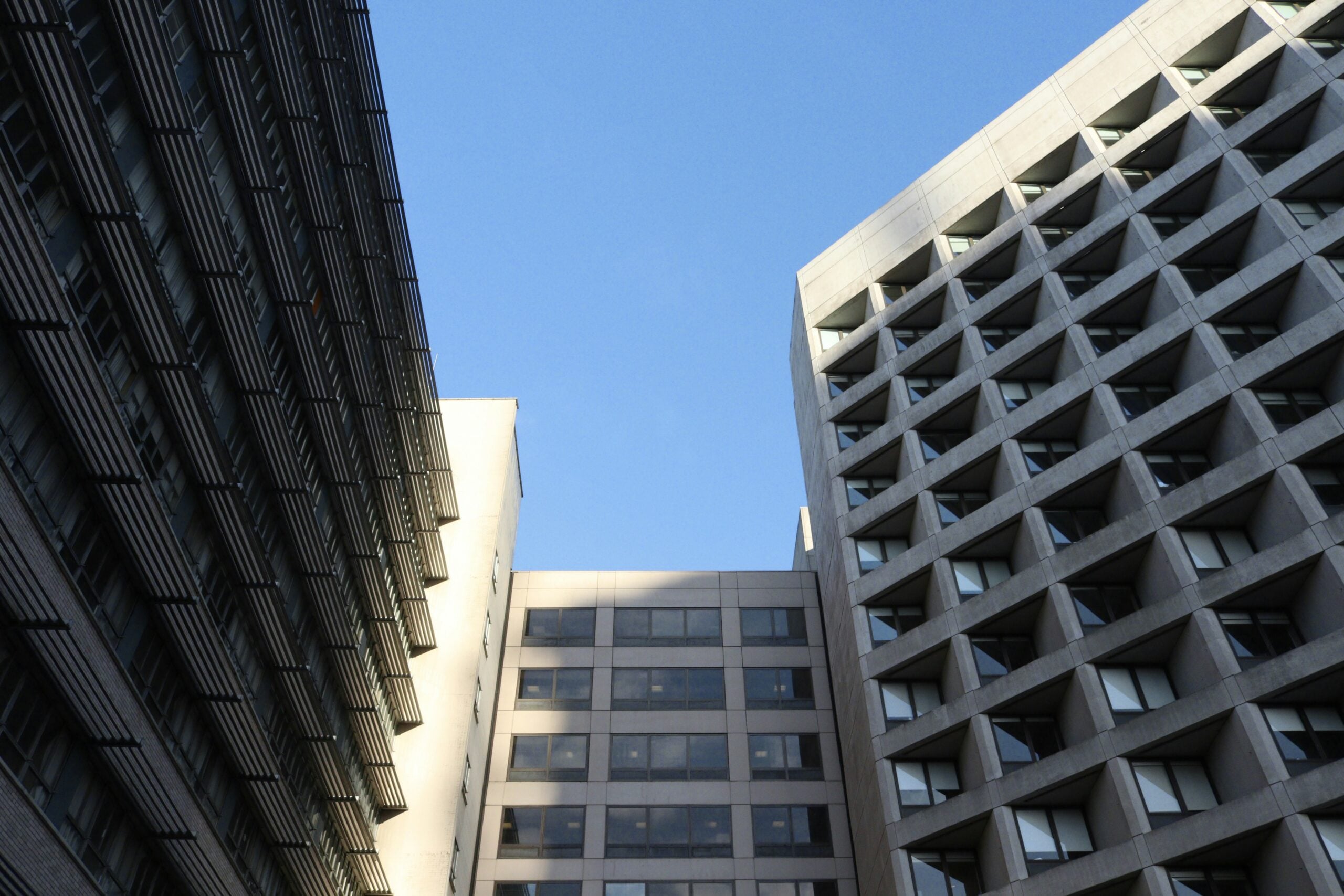 Three tall modern buildings rise toward a clear blue sky, their grid-like windows reflecting hopes for cost of living relief. Sunlight casts sharp shadows on the facades, captured from a low angle to highlight their impressive height.