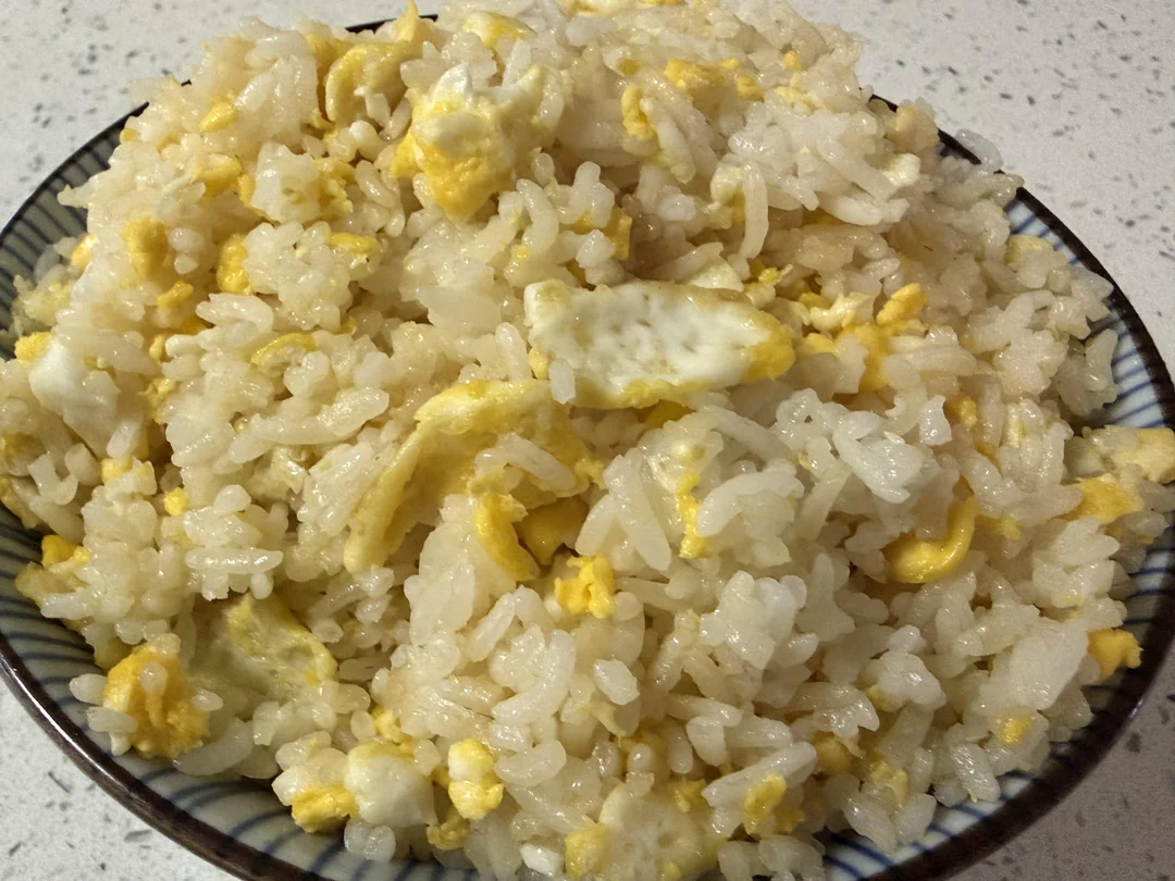 A bowl of plain fried rice with scrambled eggs, featuring white rice grains mixed with small pieces of cooked yellow egg on a speckled countertop.