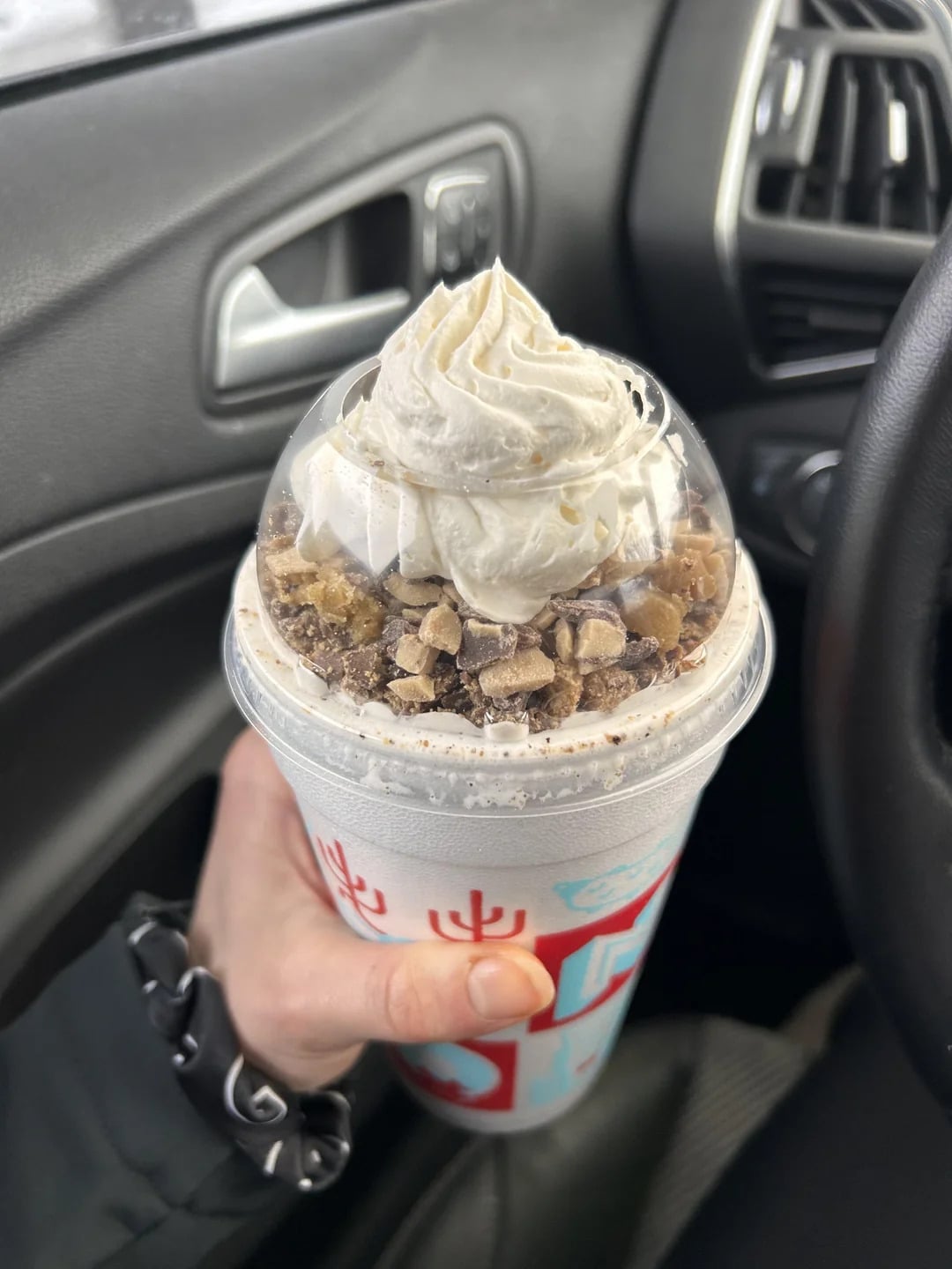 A hand holds a blended ice cream treat, one of the popular fast-food desserts, topped with whipped cream and chopped toffee bits inside a car, with a steering wheel and air vent visible in the background.