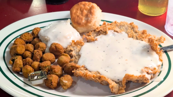A plate of chicken fried steak with white gravy, fried okra, a biscuit, and mashed potatoes topped with gravy&mdash;just what you&rsquo;d expect at the best Southern comfort food restaurants. A drink is partially visible in the background.