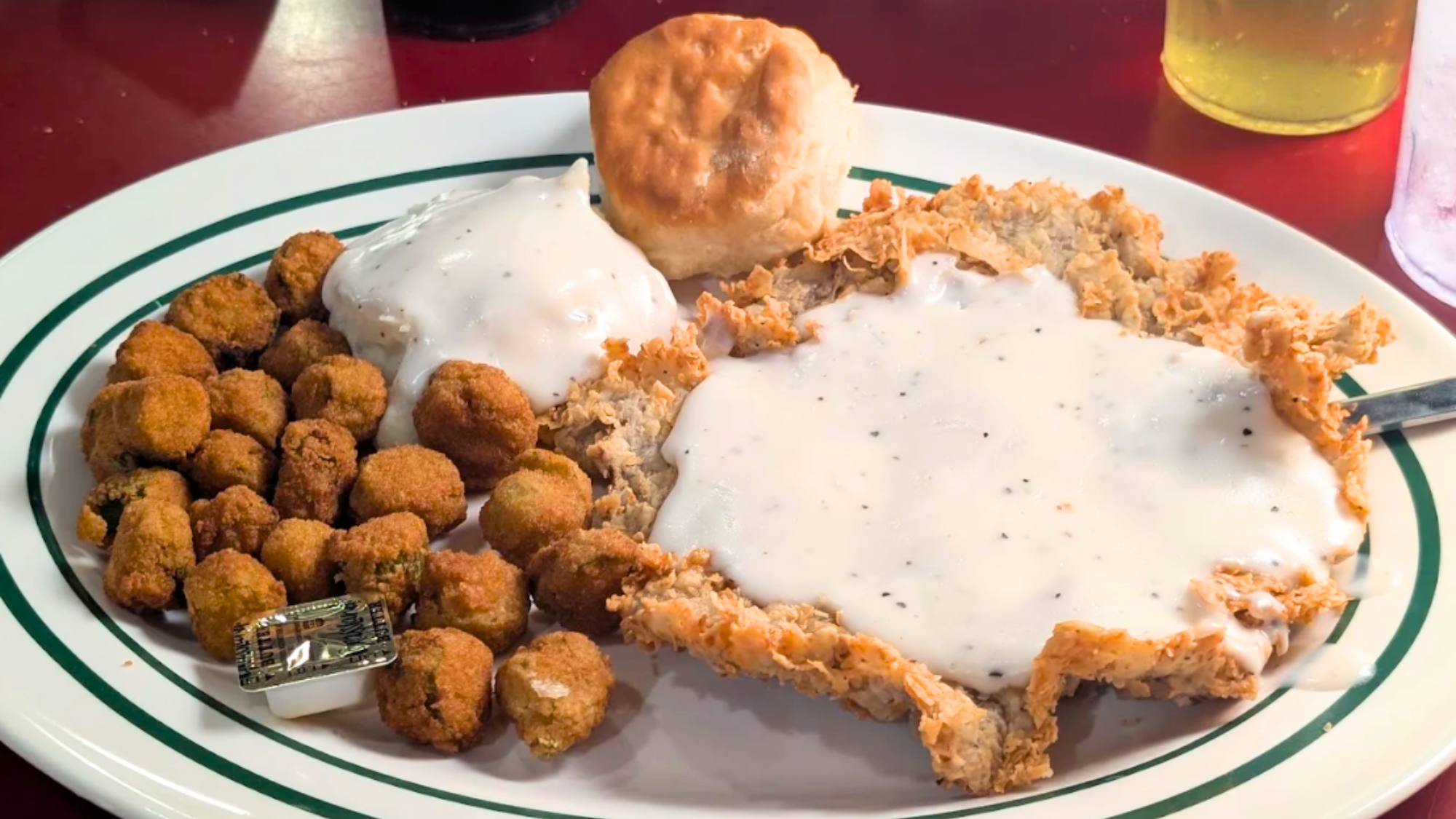 A plate of chicken fried steak with white gravy, fried okra, a biscuit, and mashed potatoes topped with gravy—just what you’d expect at the best Southern comfort food restaurants. A drink is partially visible in the background.