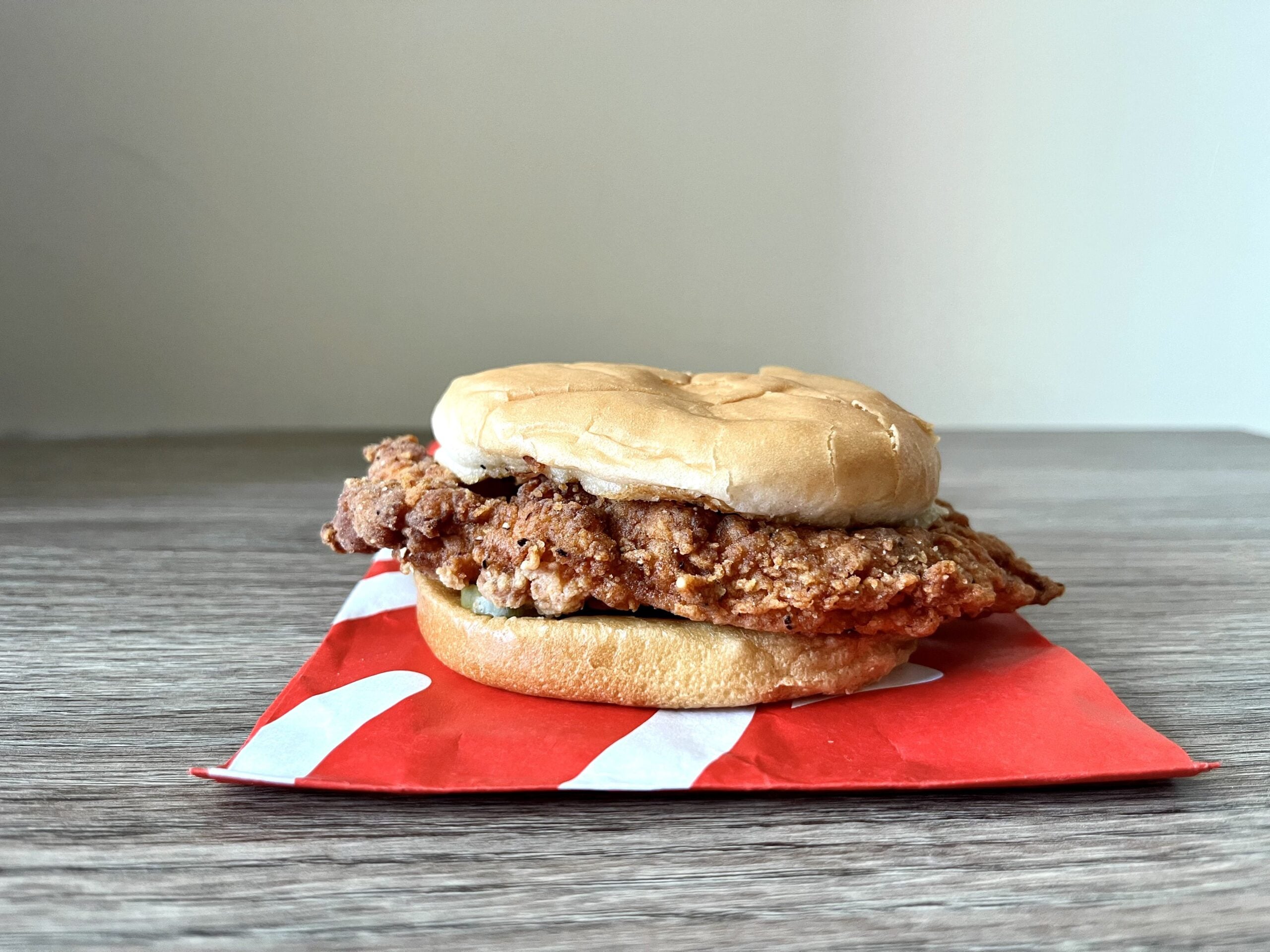 A delicious Chicken Sandwich with a crispy fried patty and soft bun sits atop a red and white paper wrapper on a wooden surface, set against a plain background.
