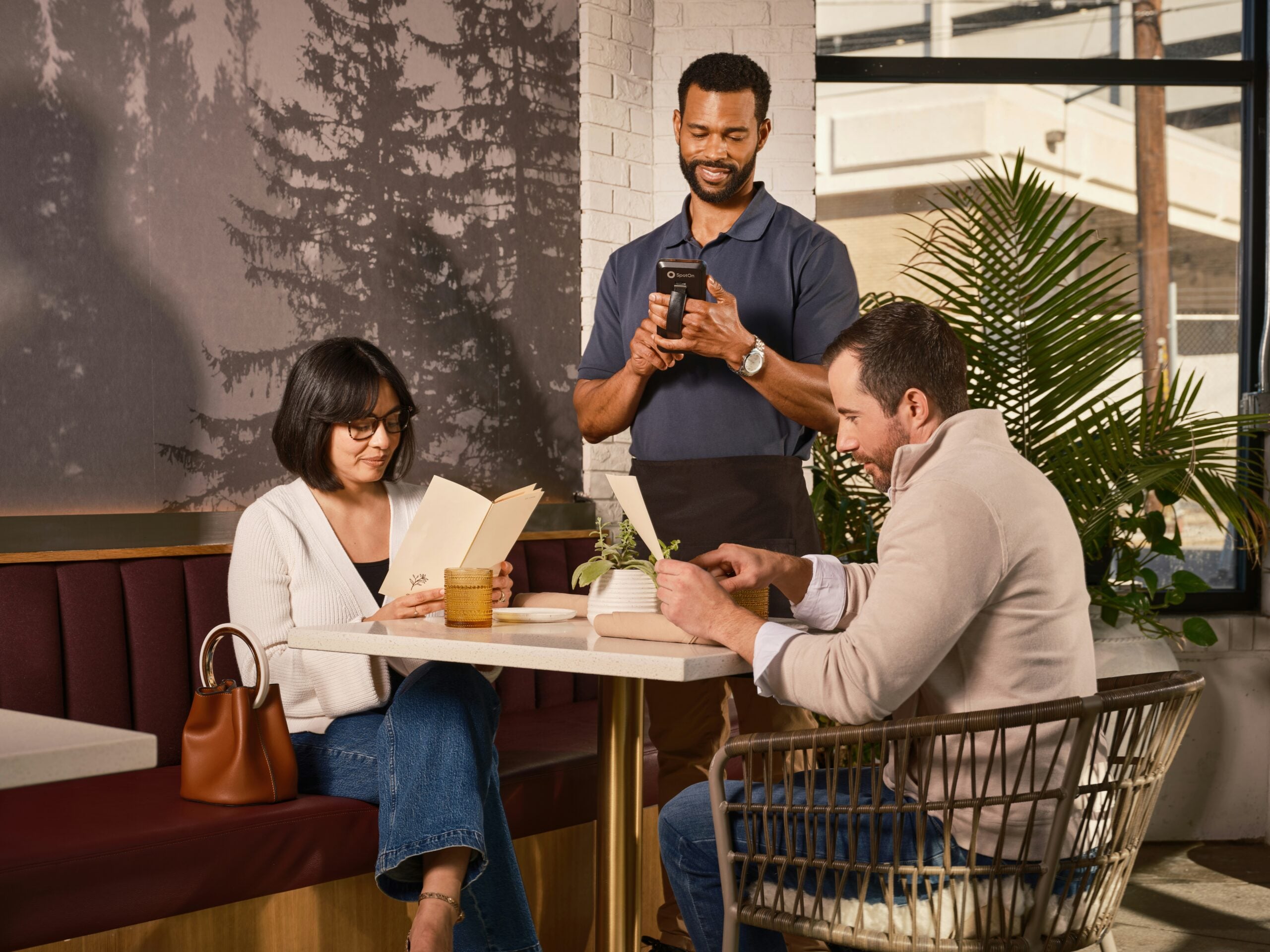 A woman and a man sit at a restaurant table looking at menus, while a smiling waiter stands nearby holding a notepad, ready to take their order. There are plants and modern decor in the background.