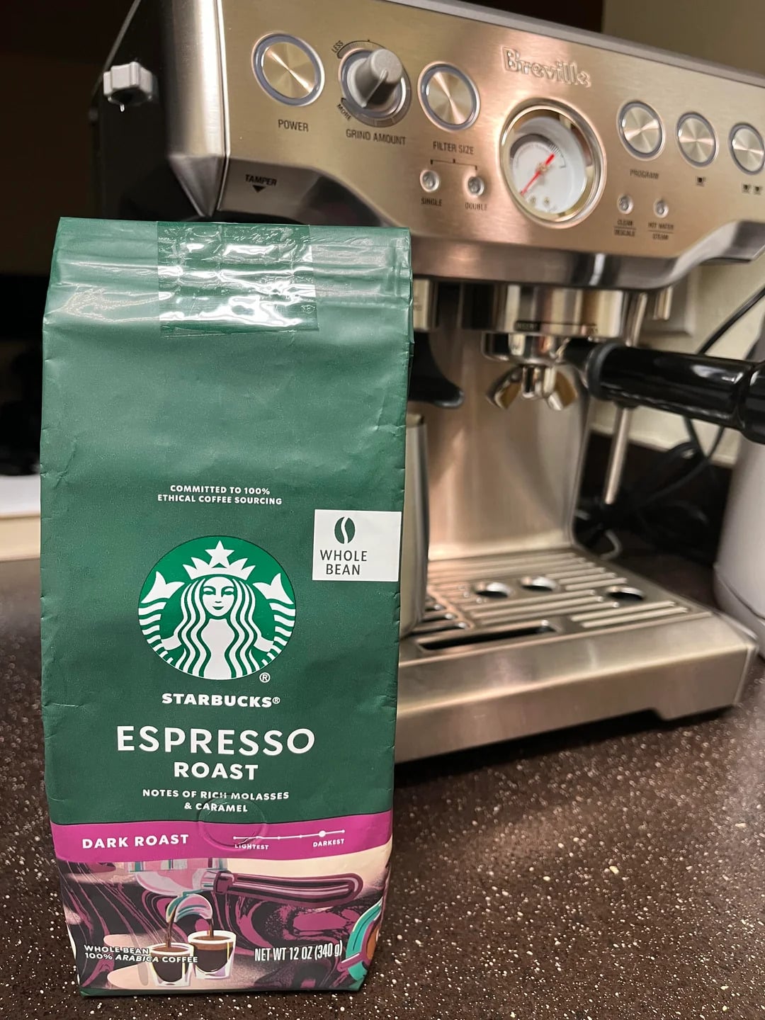 A bag of Starbucks Espresso Roast whole bean coffee, from one of the top coffee brands, sits on a kitchen counter in front of a stainless steel Breville espresso machine. The dark roast beans boast notes of rich molasses and caramel.