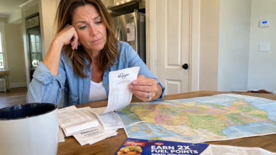 A woman sits at a table looking concerned while reviewing receipts. A map of the United States, a coffee mug, and a Kroger flyer are spread out in front of her as she searches for cost of living relief in her bright kitchen.