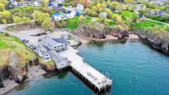 Aerial view of a coastal village with houses on green hills, a wooden pier over blue water, and rocky cliffs&mdash;an inviting spot where travelers save while enjoying breathtaking scenery.