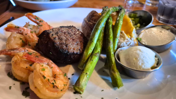 A plate from a steakhouse on the West Coast featuring grilled shrimp, filet mignon steak, roasted asparagus, a baked potato with butter and sour cream, and a side of green vegetables.