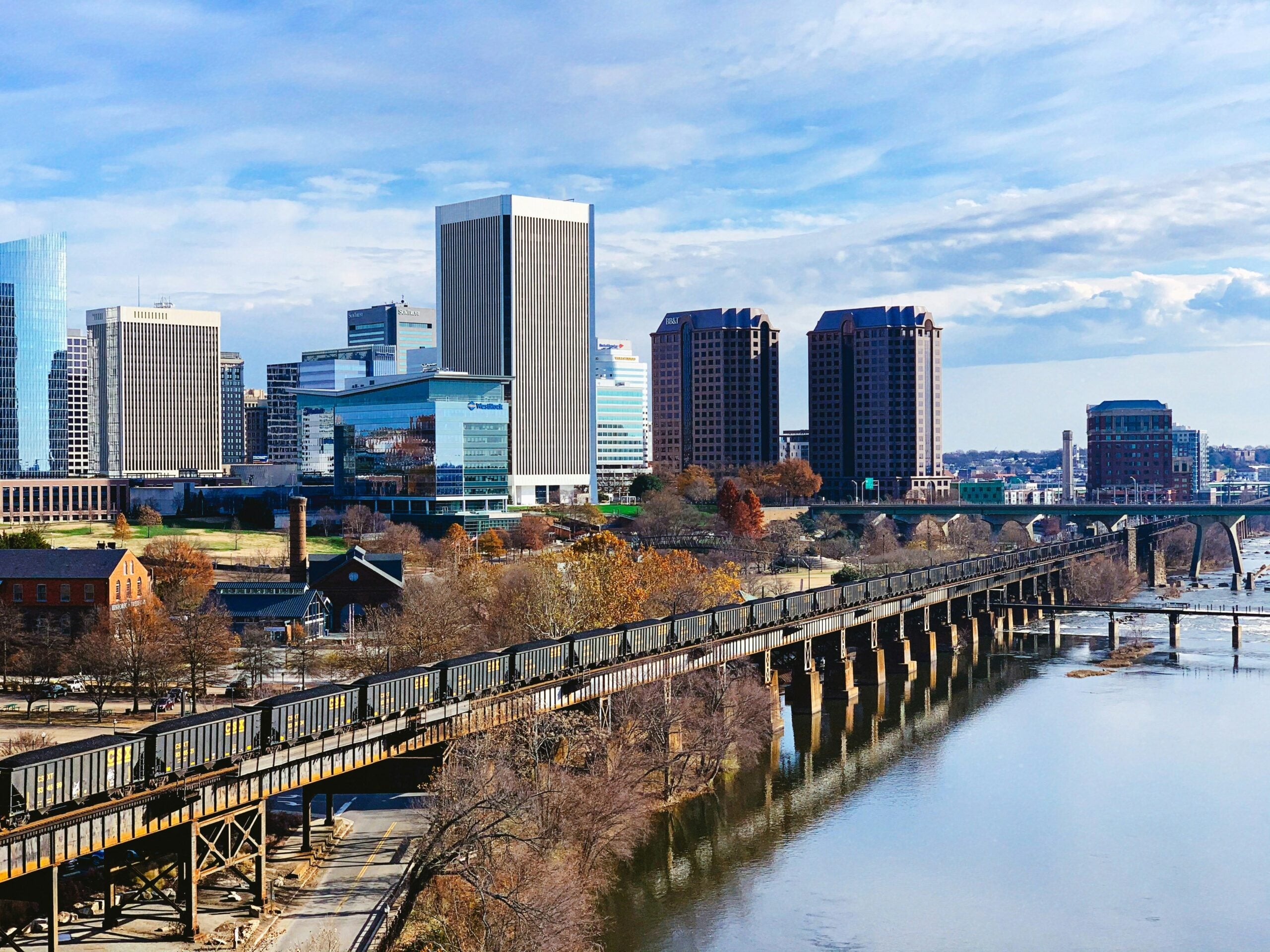 A city skyline with modern and historic buildings stands behind a river and a long railway bridge, with trees lining the water and a partly cloudy sky above.
