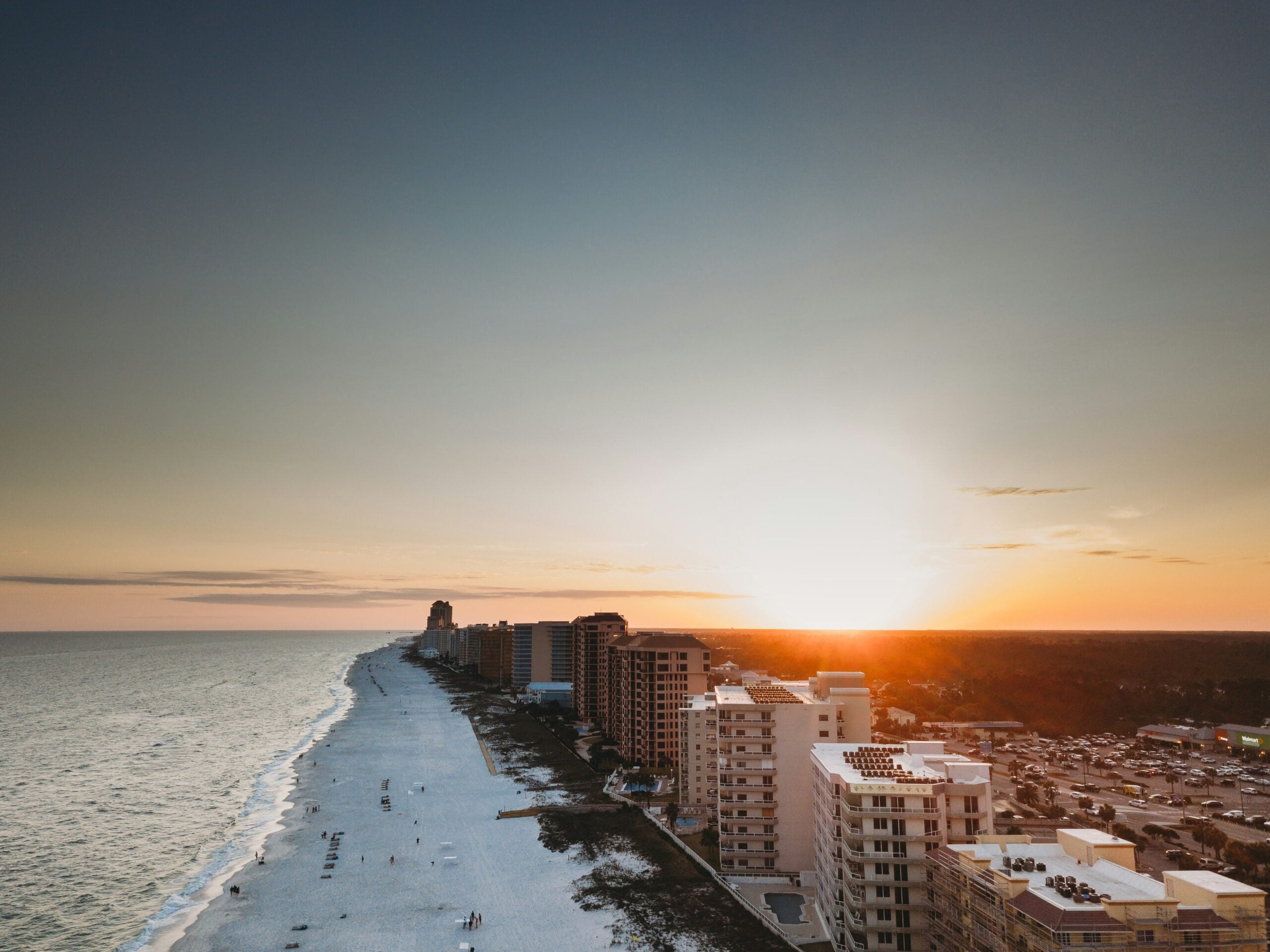 Aerial view of a coastline at sunset with high-rise buildings lining the beach, white sand, gentle waves, and a large parking area visible near the shoreline. The sun is low on the horizon, casting a warm glow.