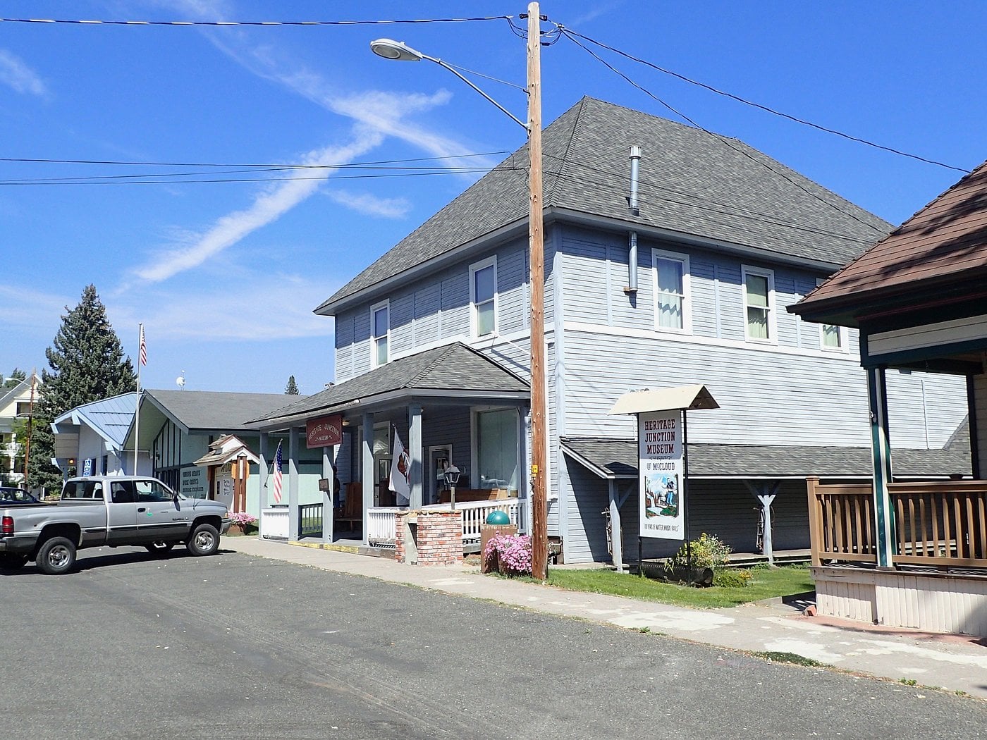 A quiet small-town street, characteristic of Unique American Towns, features a gray two-story building, a few parked vehicles, a sidewalk, and a sign out front under a clear blue sky. Power lines cross above the street.
