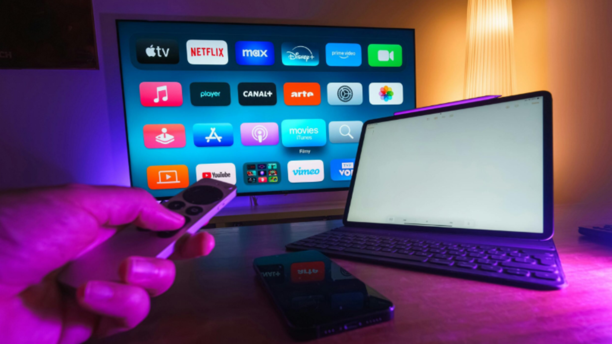 A hand holds a remote control in front of a TV displaying various streaming app icons—prompting thoughts about unused subscriptions; a tablet with a keyboard and a smartphone rest on the desk nearby, all under purple lighting.