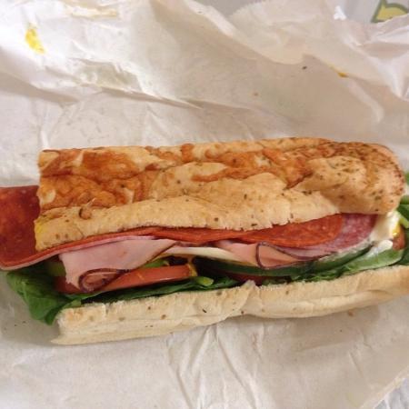 A sub sandwich on white paper, filled with layers of deli meats, cheese, lettuce, tomato, spinach, and other vegetables inside a long, toasted sub roll.