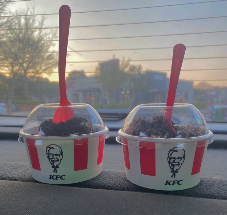 Two KFC dessert cups with red spoons and Oreo crumb toppings sit on a car dashboard, with a blurred sunset and trees visible through the rear windshield.