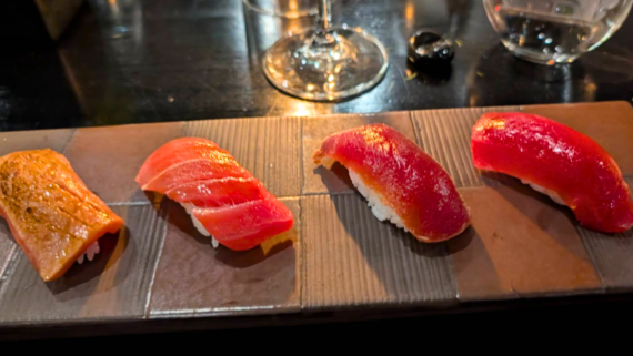 A wooden plate displays four pieces of nigiri sushi, each topped with different cuts of fish&mdash;an artful presentation you&rsquo;d expect at the best sushi restaurants. A wine glass and a water glass sit in the background on a dark table.