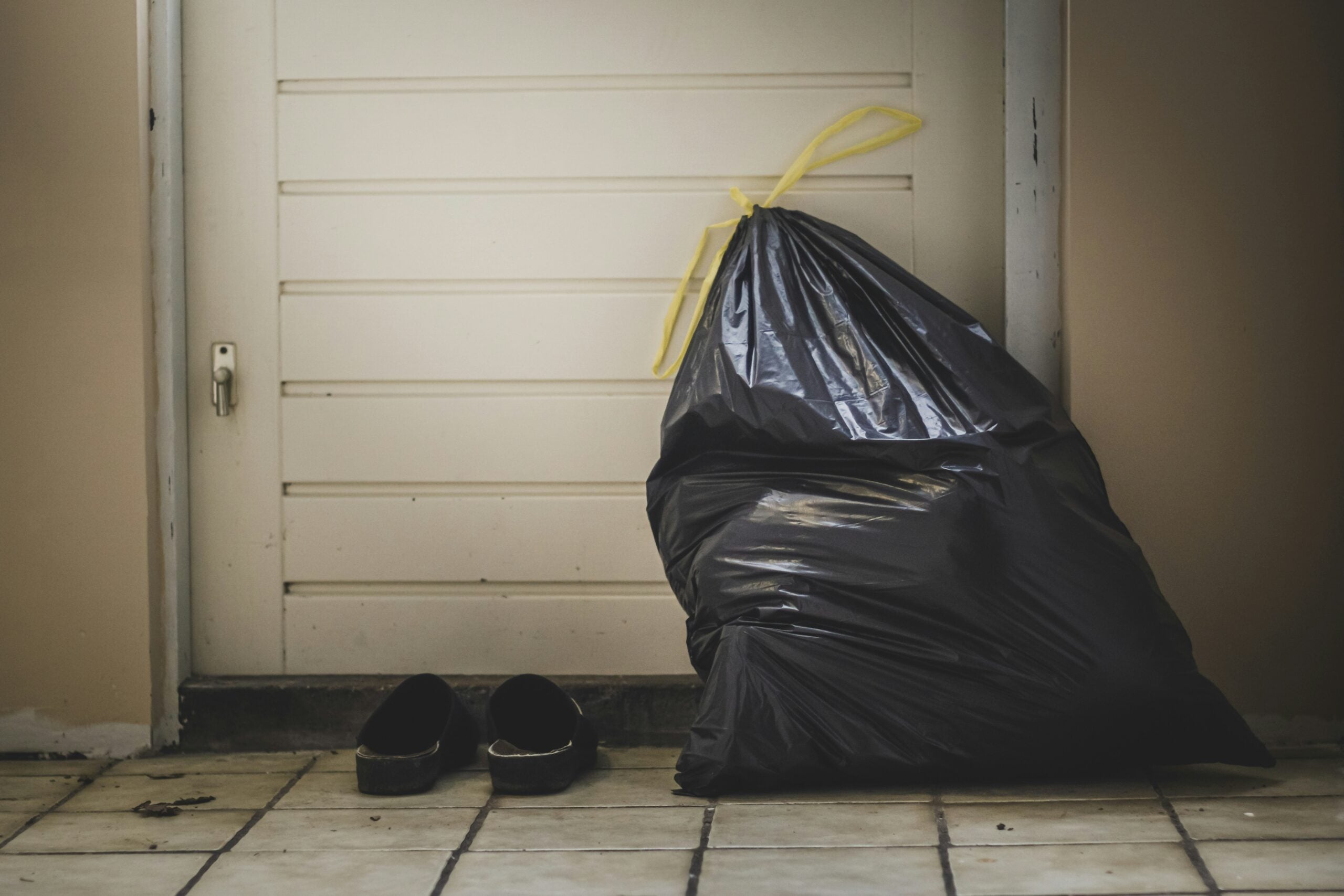 A black garbage bag tied with a yellow string sits on a tiled floor in front of a closed white door, next to a pair of black slip-on shoes.