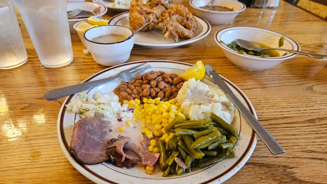 A plate of food with sliced ham, green beans, corn, mashed potatoes with gravy, baked beans, and coleslaw on a wooden table. In the background, there is a bowl of soup and a dish of fried chicken.