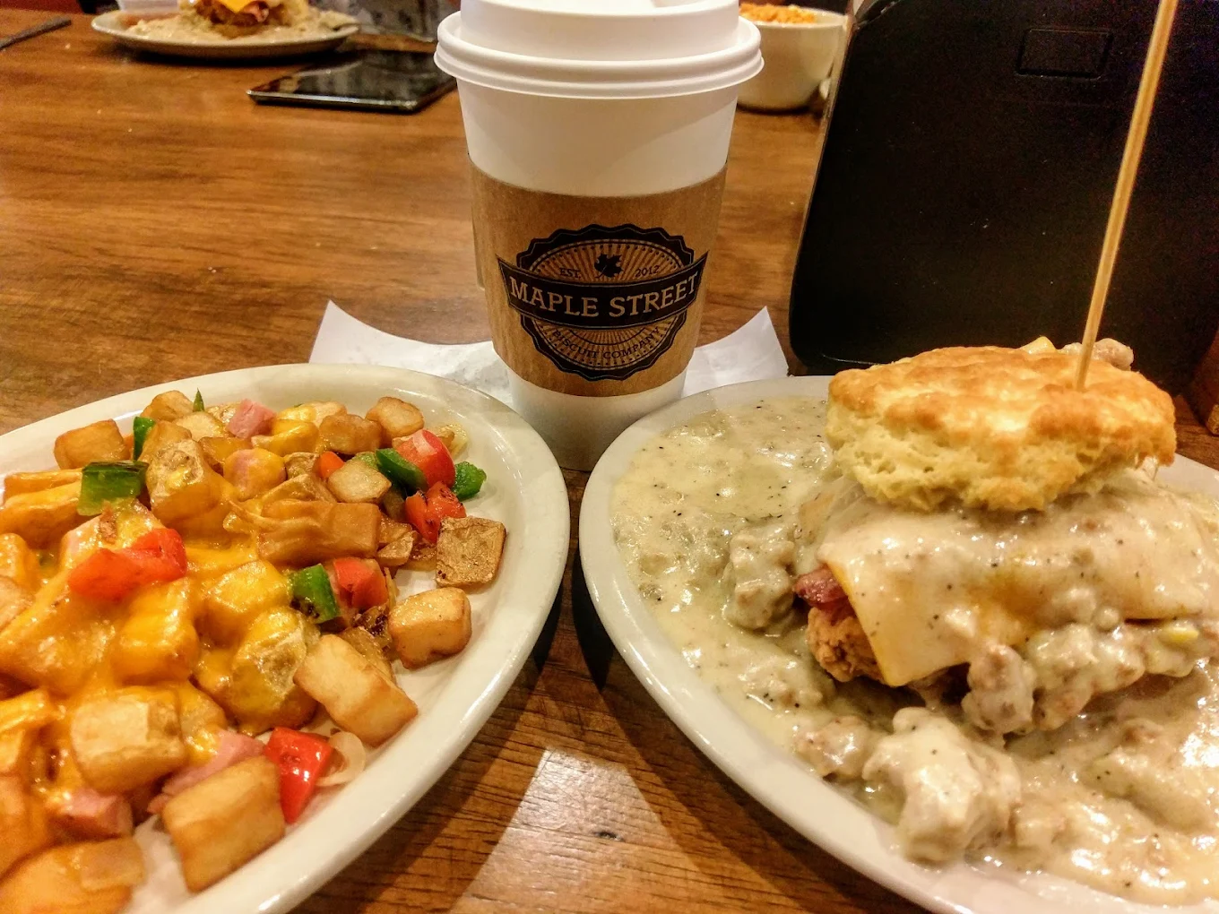 A plate of cheesy home fries with peppers, a biscuit sandwich smothered in gravy, and a coffee cup labeled "Maple Street" are arranged on a wooden table.
