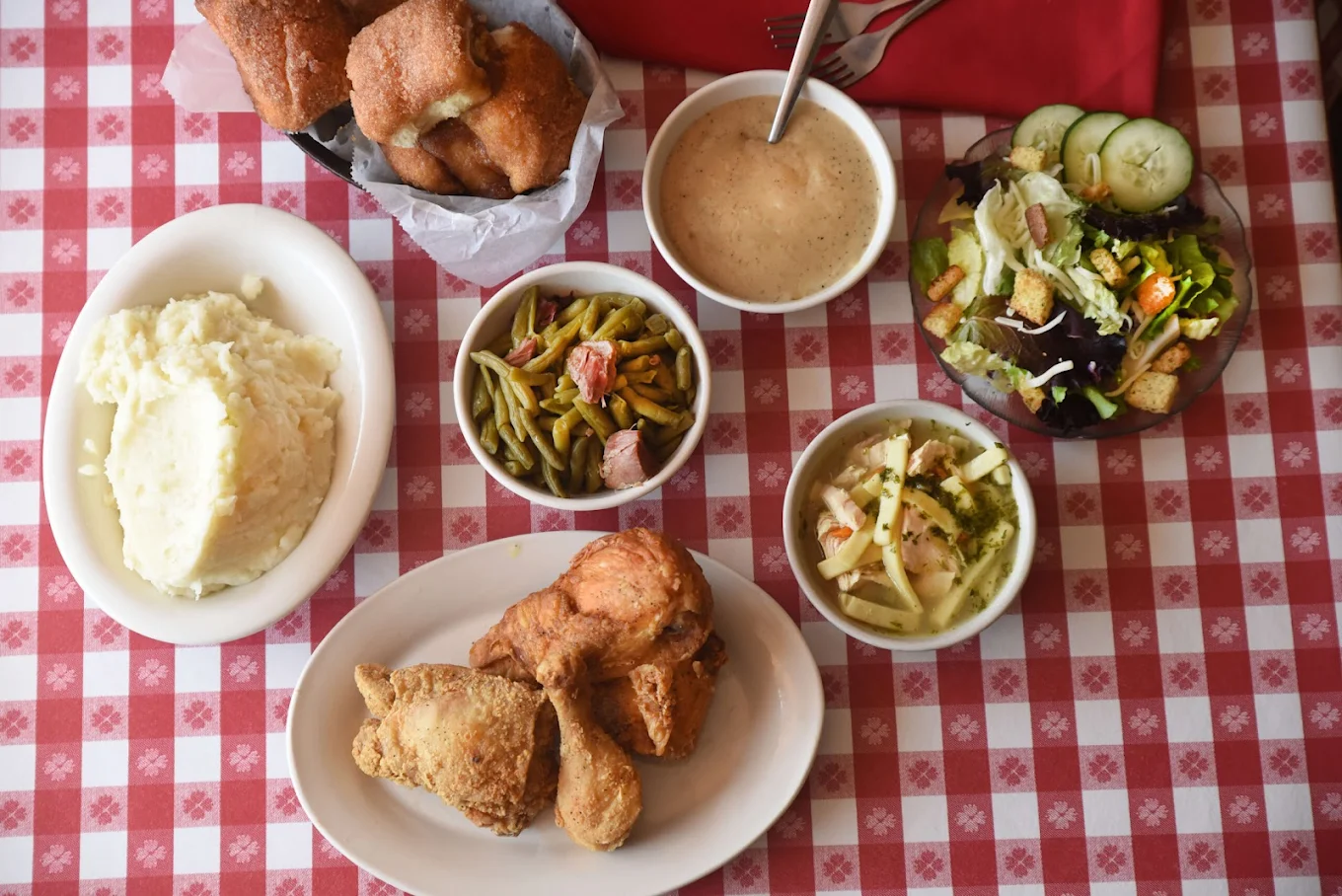 A meal on a red and white checkered tablecloth with fried chicken, mashed potatoes, green beans, coleslaw, a garden salad, rolls, and a bowl of gravy.