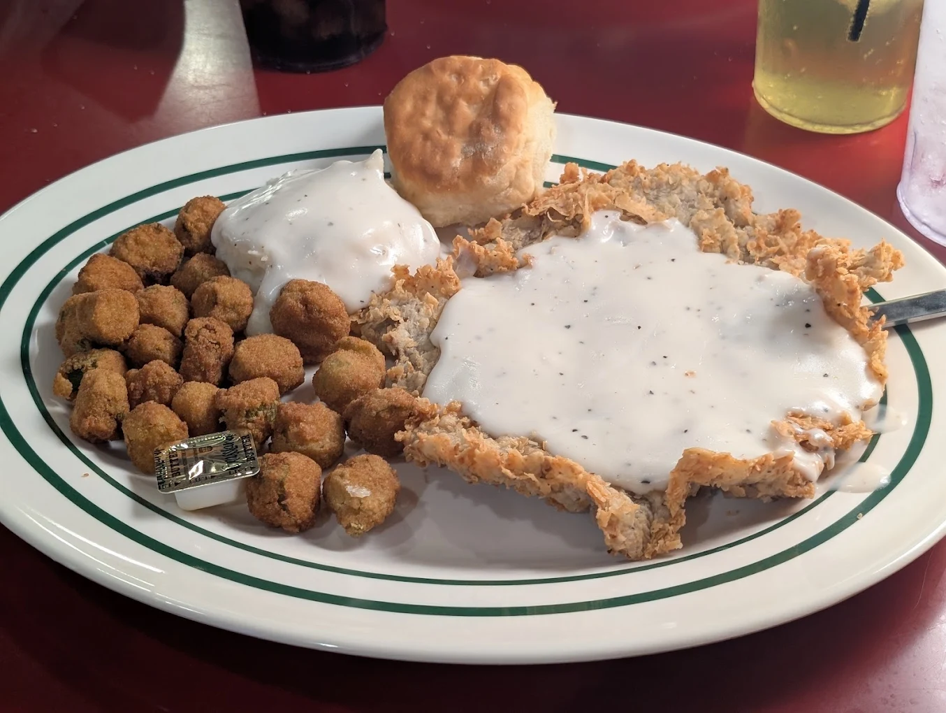 A plate of chicken fried steak topped with white gravy, fried okra, and a biscuit, served on a white plate with green stripes. Drinks are visible in the background.